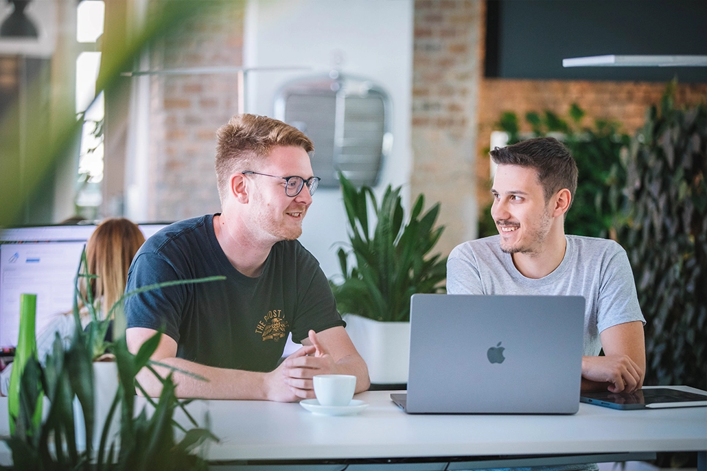Mitarbeiter*innen planen Mitarbeiterinnen und Mitarbeiter sitzen zu zweit am Laptop und tauschen sich aus.