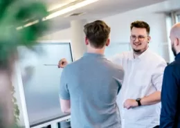 Drei Männer stehen vor einem Smartboard. Der Mann in der Mitte zeigt auf das Board.