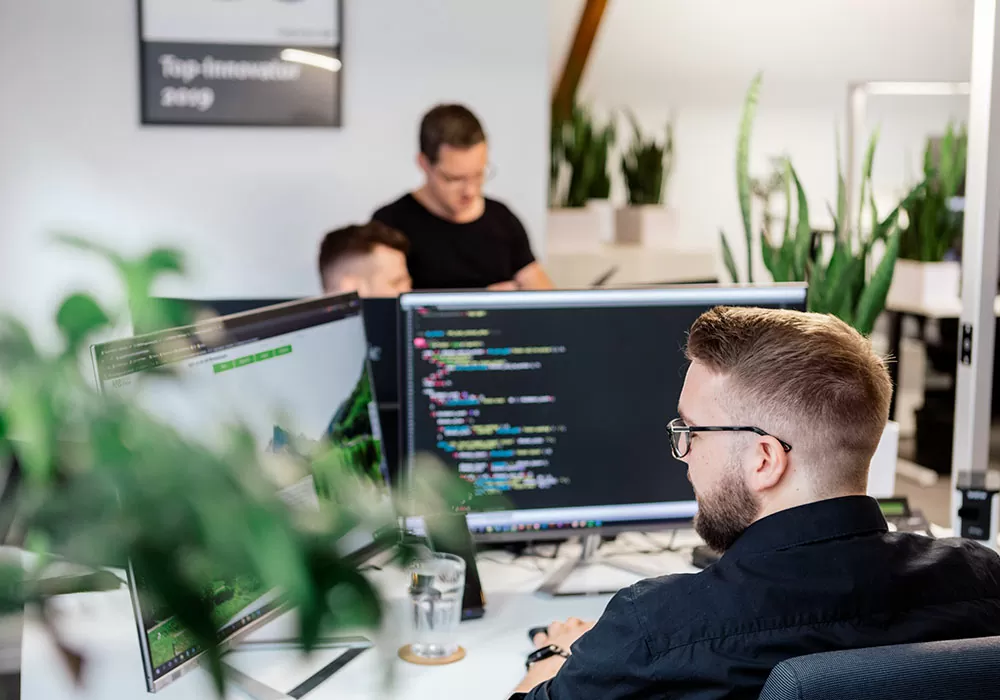 Webdesign-Agentur in Heidelberg ECONSOR Standortleiter arbeitet an seinem Arbeitsplatz. Vor ihm stehen 2 Bildschirme. Im Hintergrund sieht man zwei weitere Mitarbeiter miteinander reden.