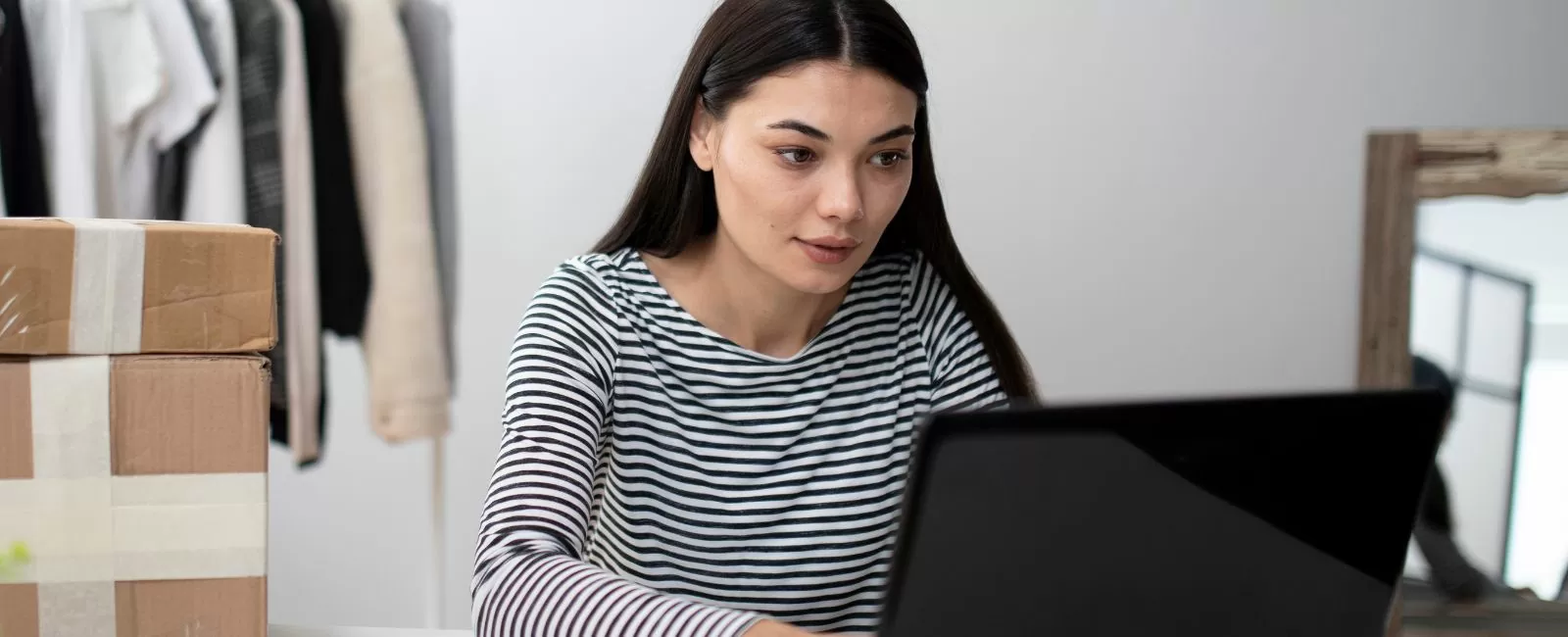 Frau sitzt am PC umgeben von Kartons und Kleidung